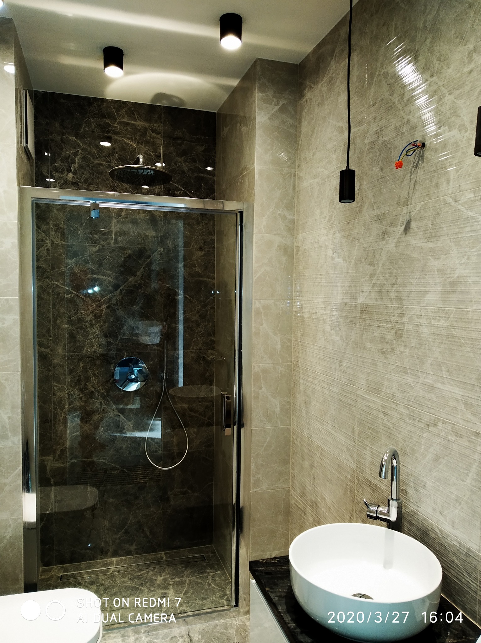 Nowoczesna łazienka z kabiną prysznicową typu walk-in, wykończona marmurowymi płytkami w odcieniach szarości i brązu, oświetlona punktowymi lampami sufitowymi i wiszącą lampą z odsłoniętymi...