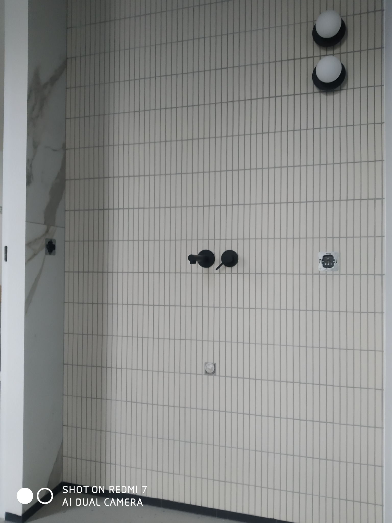 Ściana w łazience w Gdańsku wyłożona pionowymi, wąskimi, beżowymi płytkami, z widocznymi czarnymi bateriami, okrągłymi lampami i gniazdkiem elektrycznym, w trakcie montażu.