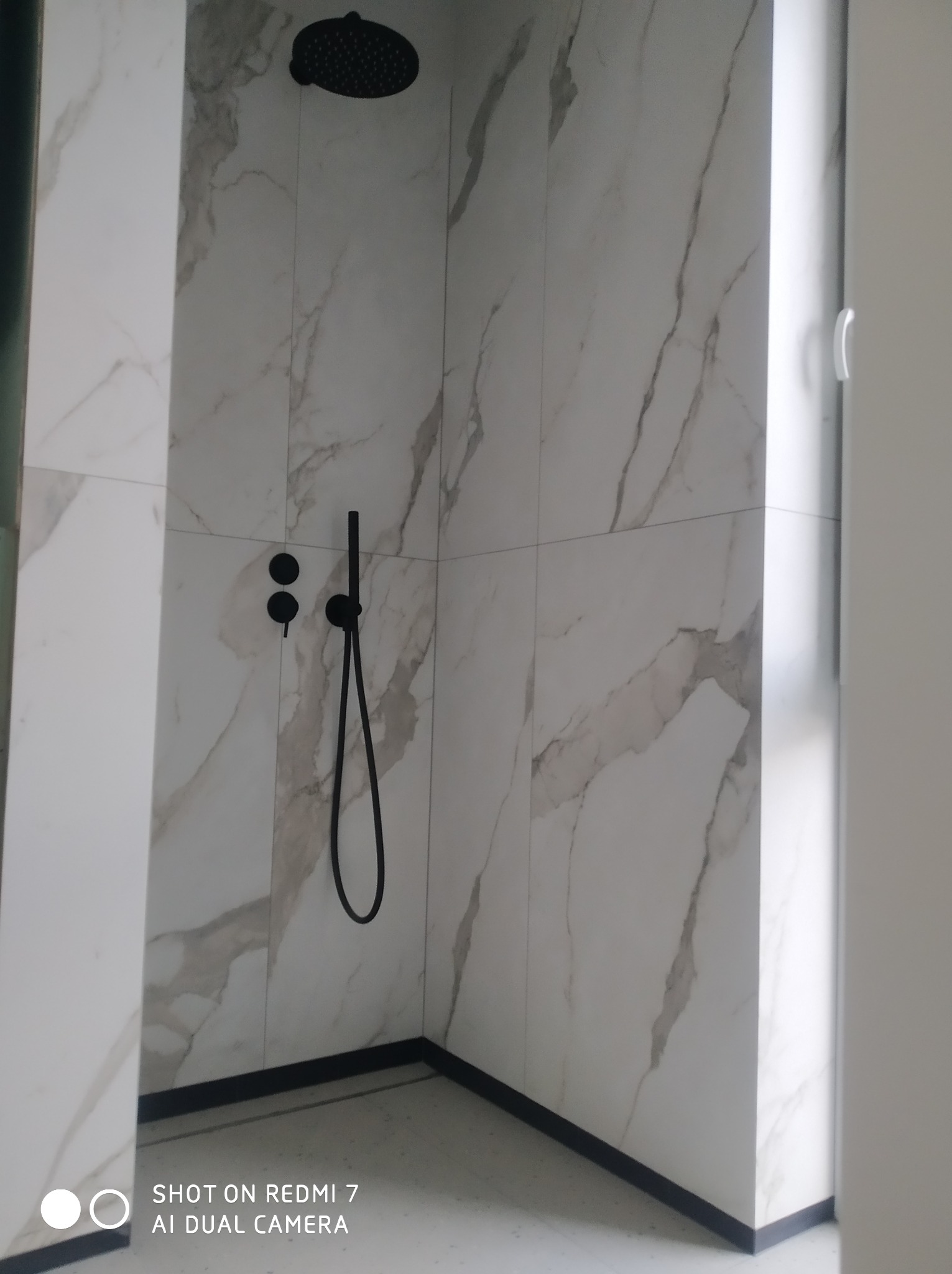 Nowoczesna kabina prysznicowa wykończona białymi płytkami imitującymi marmur, z czarną armaturą i deszczownicą, widoczna z perspektywy wejścia do łazienki.