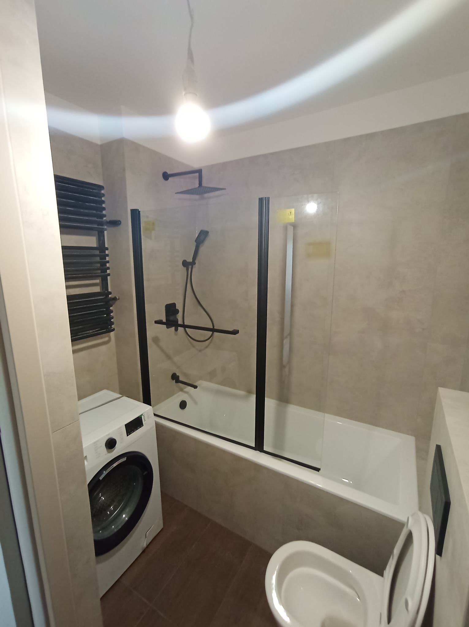 Nowoczesna łazienka z beżowymi płytkami, czarnymi akcentami w postaci kabiny prysznicowej, grzejnika i armatury, białą wanną, pralką i toaletą.