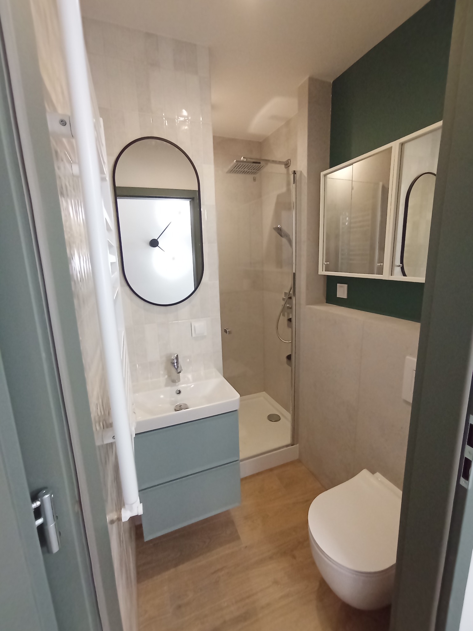 Nowoczesna łazienka z umywalką na szafce w kolorze pastelowej zieleni, owalnym lustrem z czarną ramą-zegarem, kabiną prysznicową i toaletą w zabudowie, wykończona płytkami w odcieniach bieli i beżu...