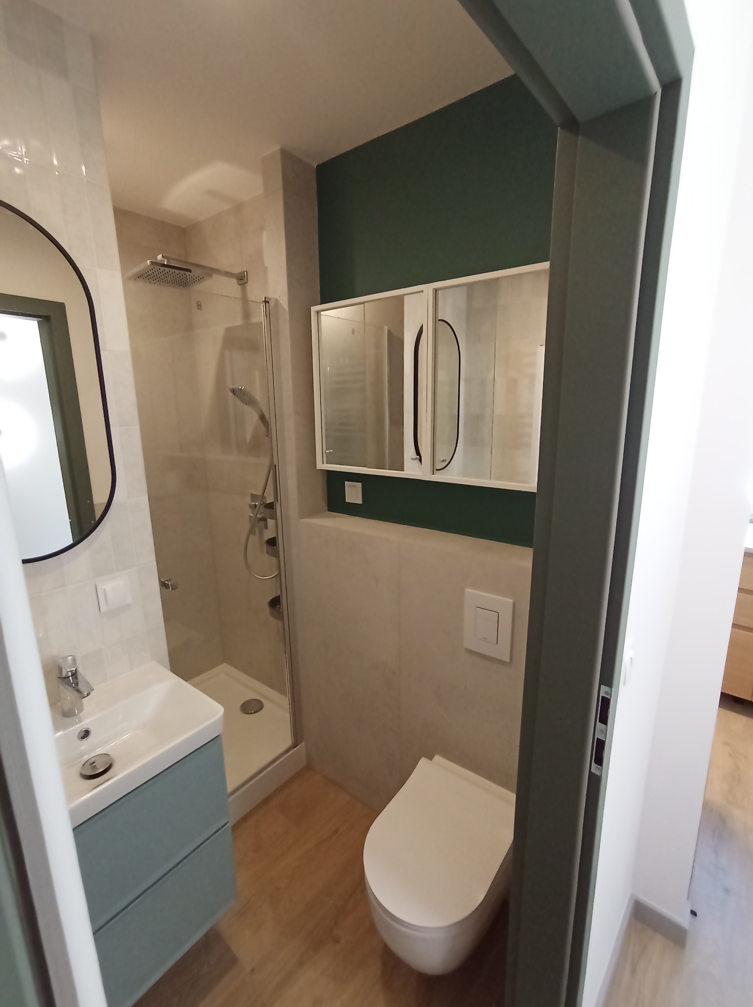 Nowoczesna łazienka z kabiną prysznicową typu walk-in, umywalką z szafką w kolorze morskim i wiszącą toaletą, wykończona płytkami w odcieniach beżu i zieleni, widoczna z perspektywy wejścia.