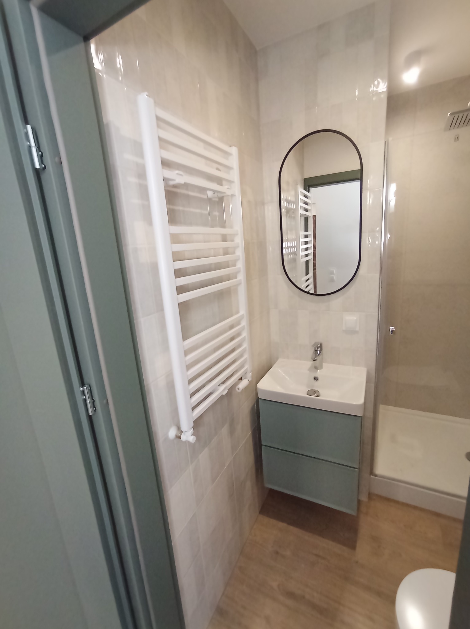 Ukończona łazienka z kabiną prysznicową, umywalką z szafką w kolorze morskim, grzejnikiem drabinkowym i owalnym lustrem z czarną ramą, widoczna przez otwarte drzwi