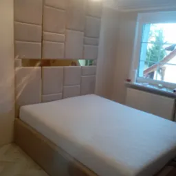 Beżowe łóżko z wezgłowiem z paneli tapicerowanych w różnych rozmiarach, przedzielonych poziomym pasem lustra, w tle okno.