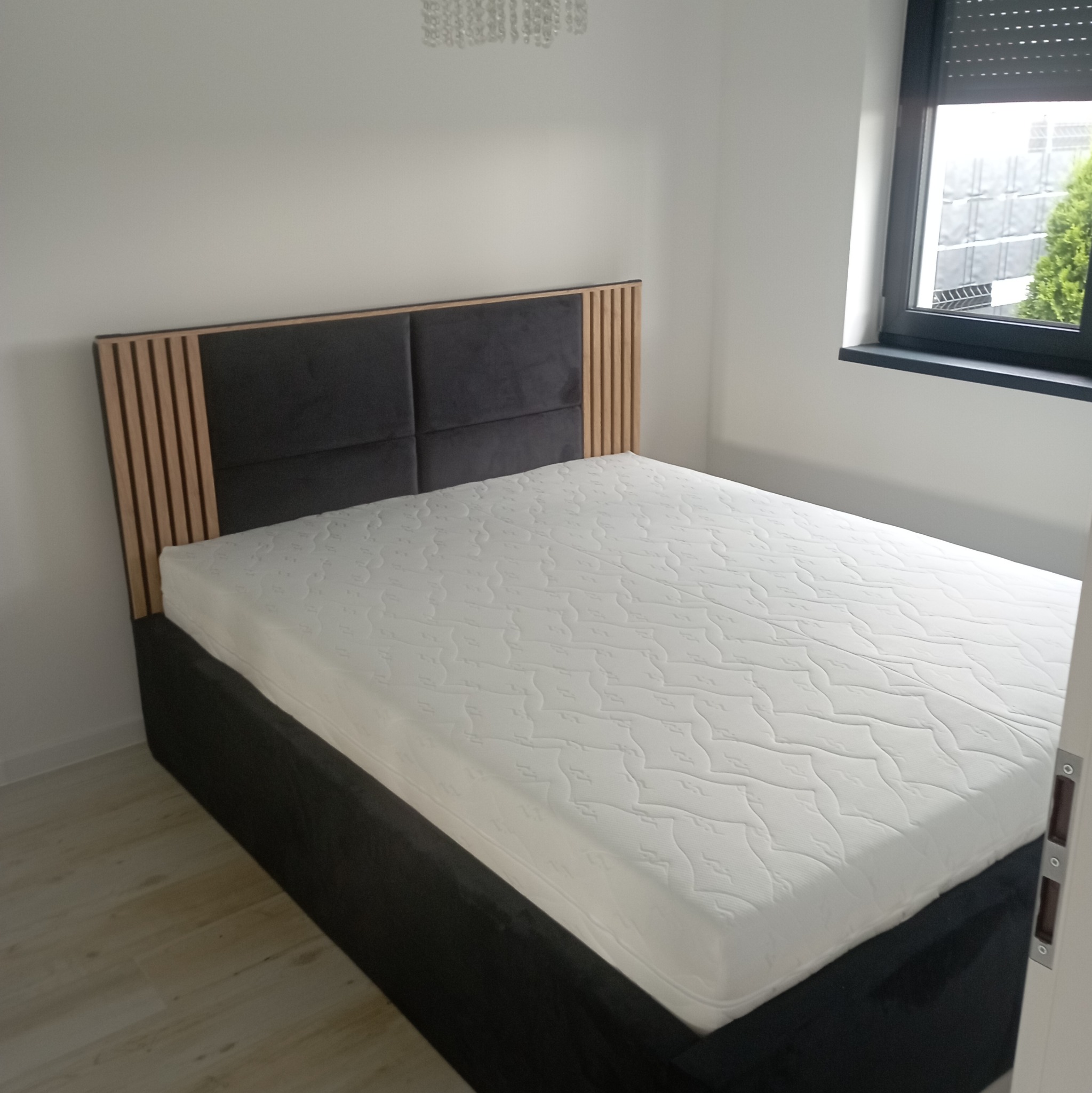Łóżko z tapicerowanym, ciemnoszarym wezgłowiem z drewnianymi listwami i białym materacem w minimalistycznej sypialni z oknem.