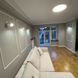 AKA STUDIO - Jasnoszary salon po remoncie z parkietem w jodełkę, ozdobnymi listwami na ścianach i nowoczesnym oświetleniem.