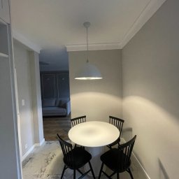 AKA STUDIO - Jasne wnętrze jadalni z okrągłym białym stołem, czterema czarnymi krzesłami i wiszącą lampą. W tle widoczny salon z szarą sofą i dekoracyjnymi panelami ściennymi. Ściany pomalowane na jasny odcień...