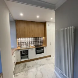 Wykończona kuchnia z białymi szafkami dolnymi i drewnianymi górnymi, wzorzysta glazura nad blatem, marmurowa podłoga i biały grzejnik żeberkowy.