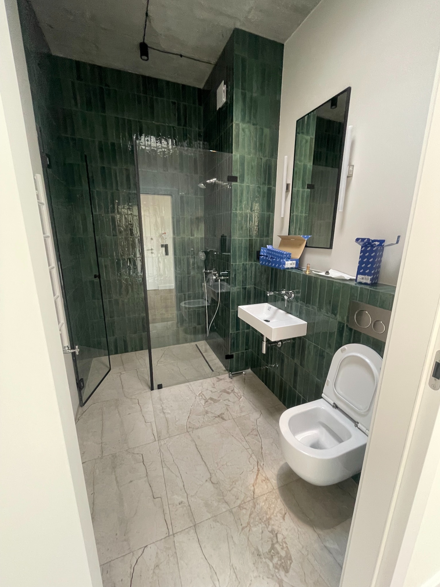 Nowoczesna łazienka z kabiną prysznicową typu walk-in, wykończona ciemnozielonymi, pionowo ułożonymi płytkami, umywalką nablatową i wiszącą toaletą. Na blacie widoczne opakowania armatury...