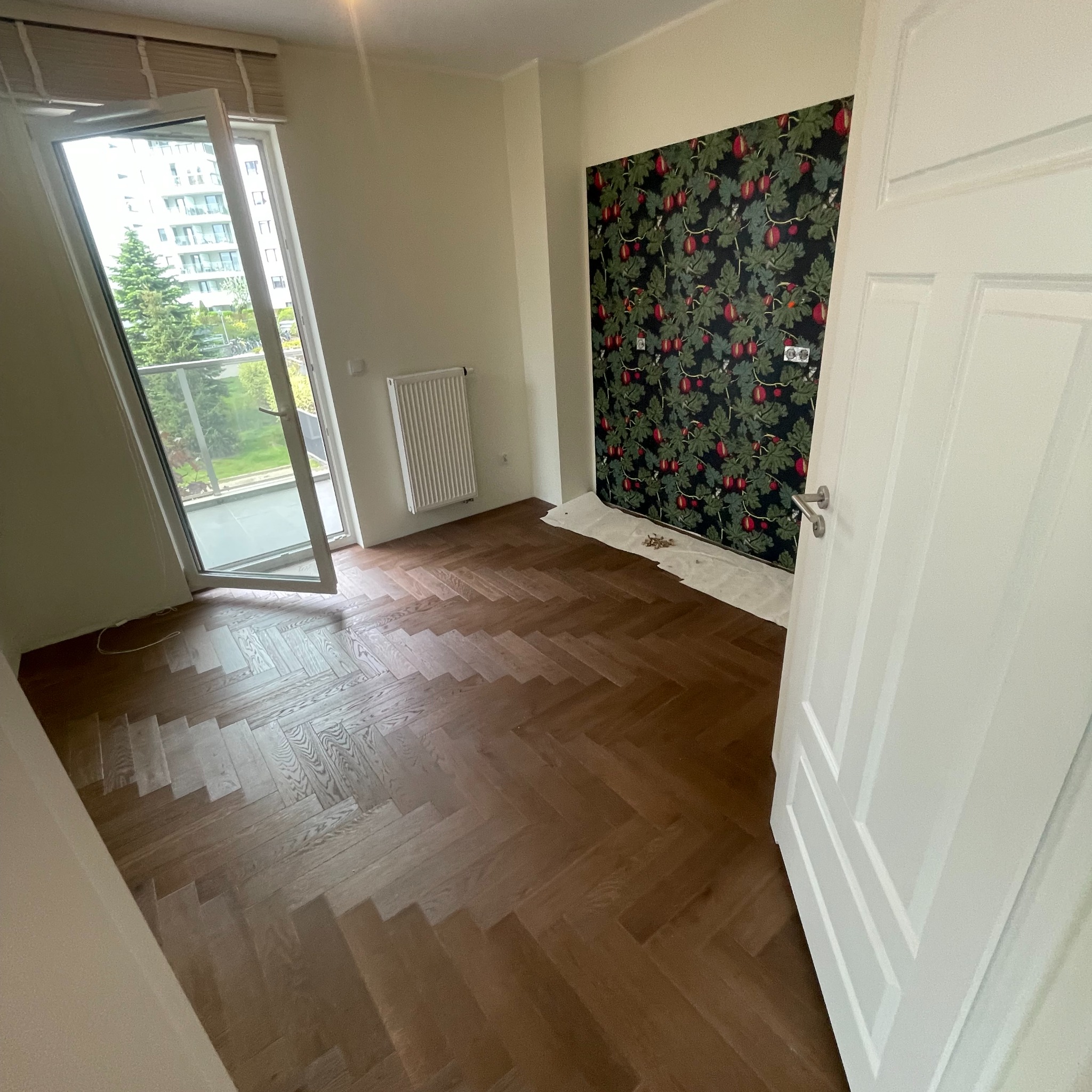 Pokój w trakcie remontu: ułożona drewniana podłoga w jodełkę, ściana z fragmentem tapety w motywy roślinne, otwarte drzwi balkonowe z widokiem na zieleń.