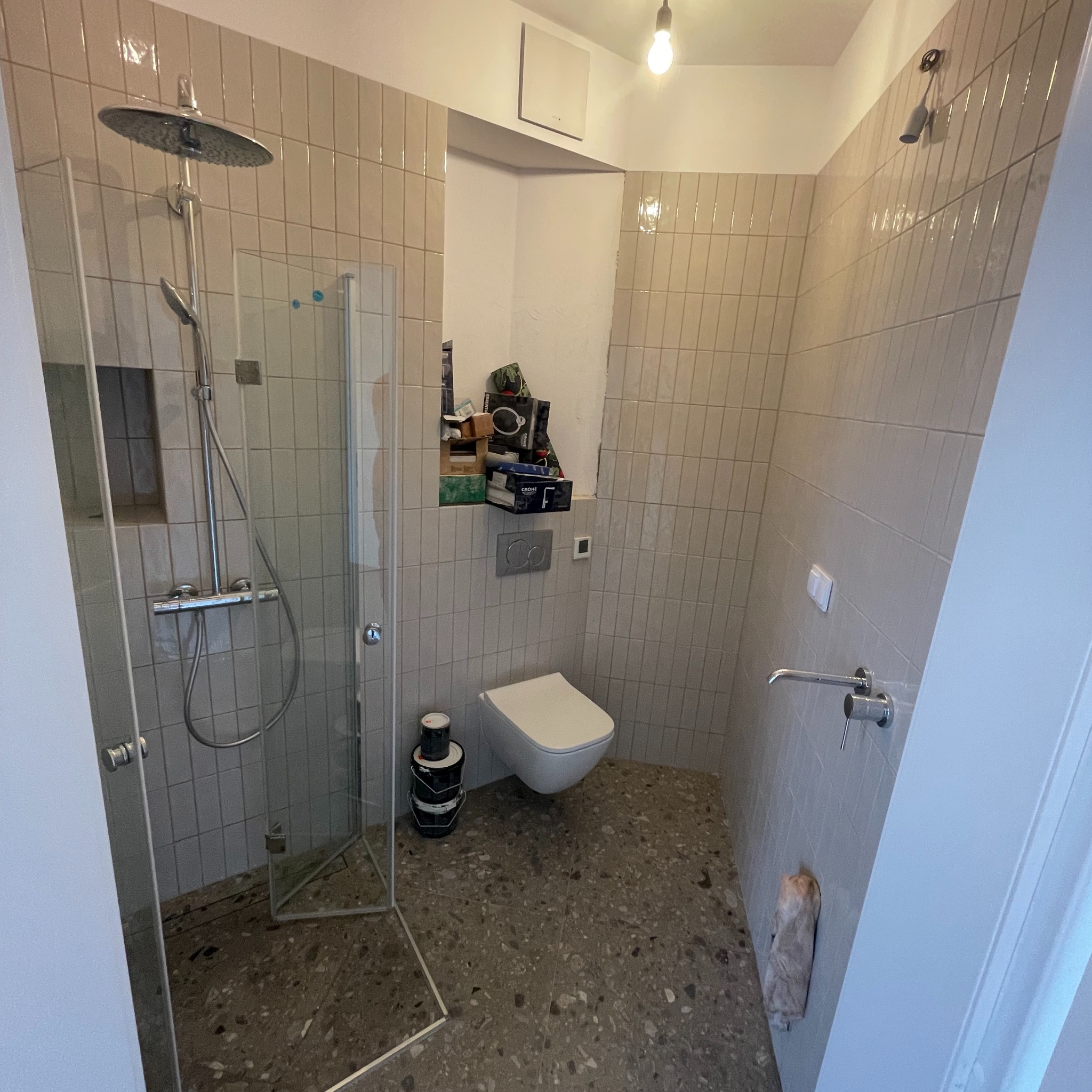 Ukończona łazienka z kabiną prysznicową typu walk-in, toaletą wiszącą, szarą mozaiką na podłodze i ścianach wyłożonych pionowo ułożonymi płytkami w odcieniach beżu.