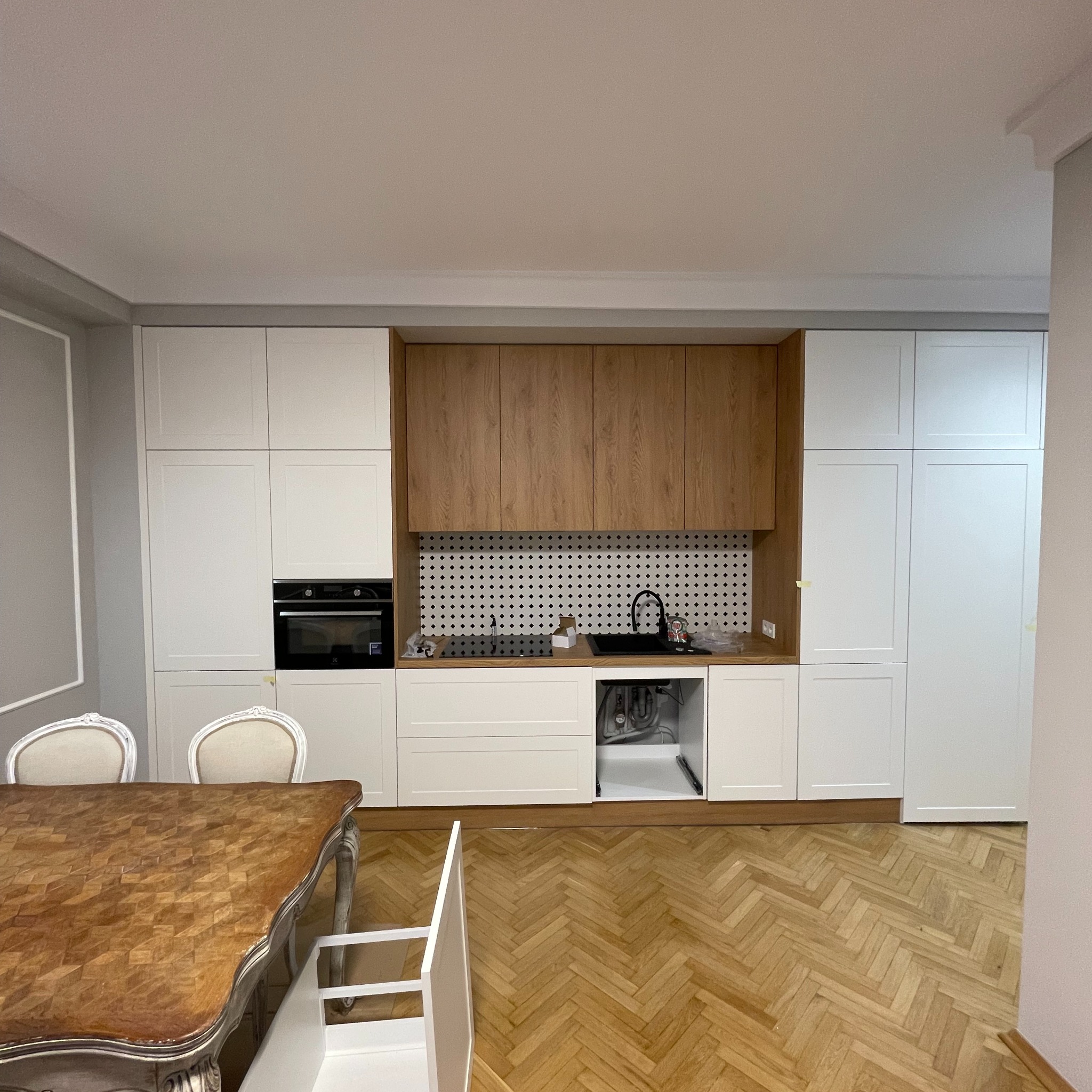 Nowoczesna kuchnia z białymi i drewnianymi szafkami, czarnym zlewem i płytą indukcyjną, widoczne instalacje pod zlewem, parkiet w jodełkę na podłodze, stół z krzesłami w stylu vintage.