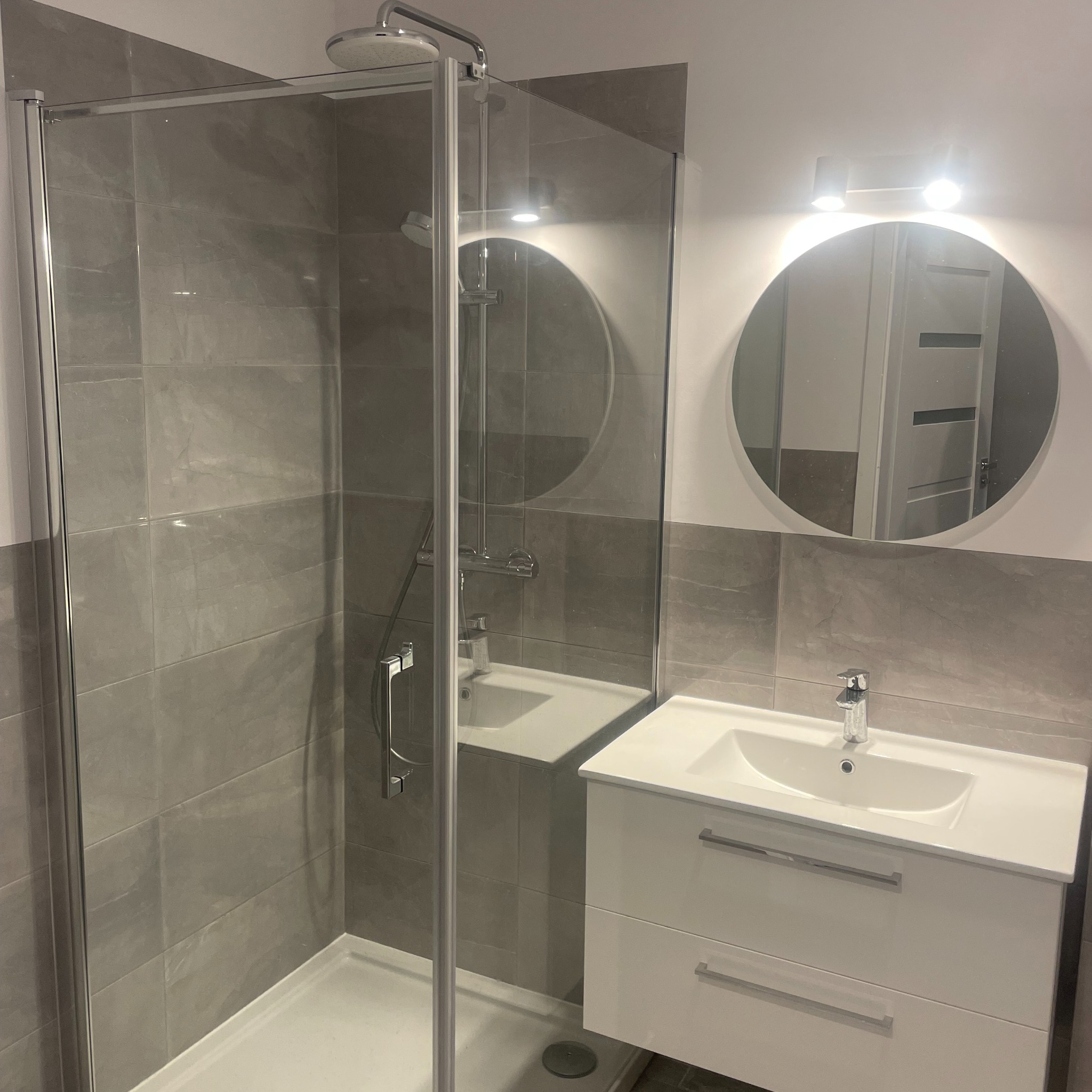 Nowoczesna łazienka z kabiną prysznicową typu walk-in, szarą glazurą imitującą kamień i białą szafką z umywalką oraz okrągłym lustrem podświetlanym LED.