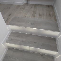 Rida Zarzeczny MASTER BUD - Schody wykończone panelami imitującymi drewno w Garbatce-Letnisko, z widoczną listwą wykończeniową i grzejnikiem na podłodze.