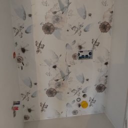 Rida Zarzeczny MASTER BUD - Świeżo wykafelkowana ściana łazienki z motywem kwiatowym, widoczne przygotowane punkty instalacji wodnej i kanalizacyjnej, żółta osłona na rurę odpływową.