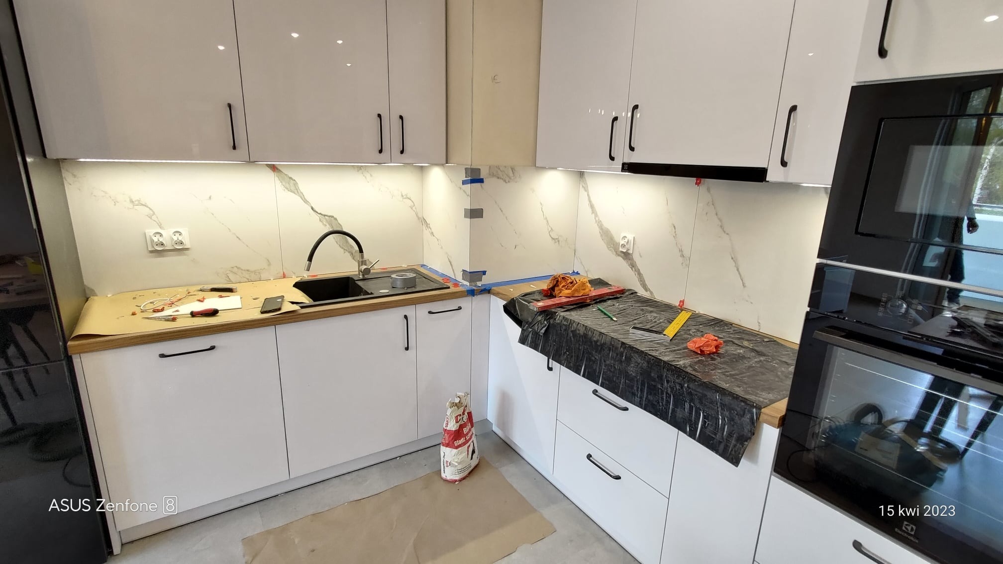Biała kuchnia z marmurowym wzorem na ścianie w trakcie montażu płytek, widoczne narzędzia, poziomica, worek z zaprawą klejową i zabezpieczone blaty.