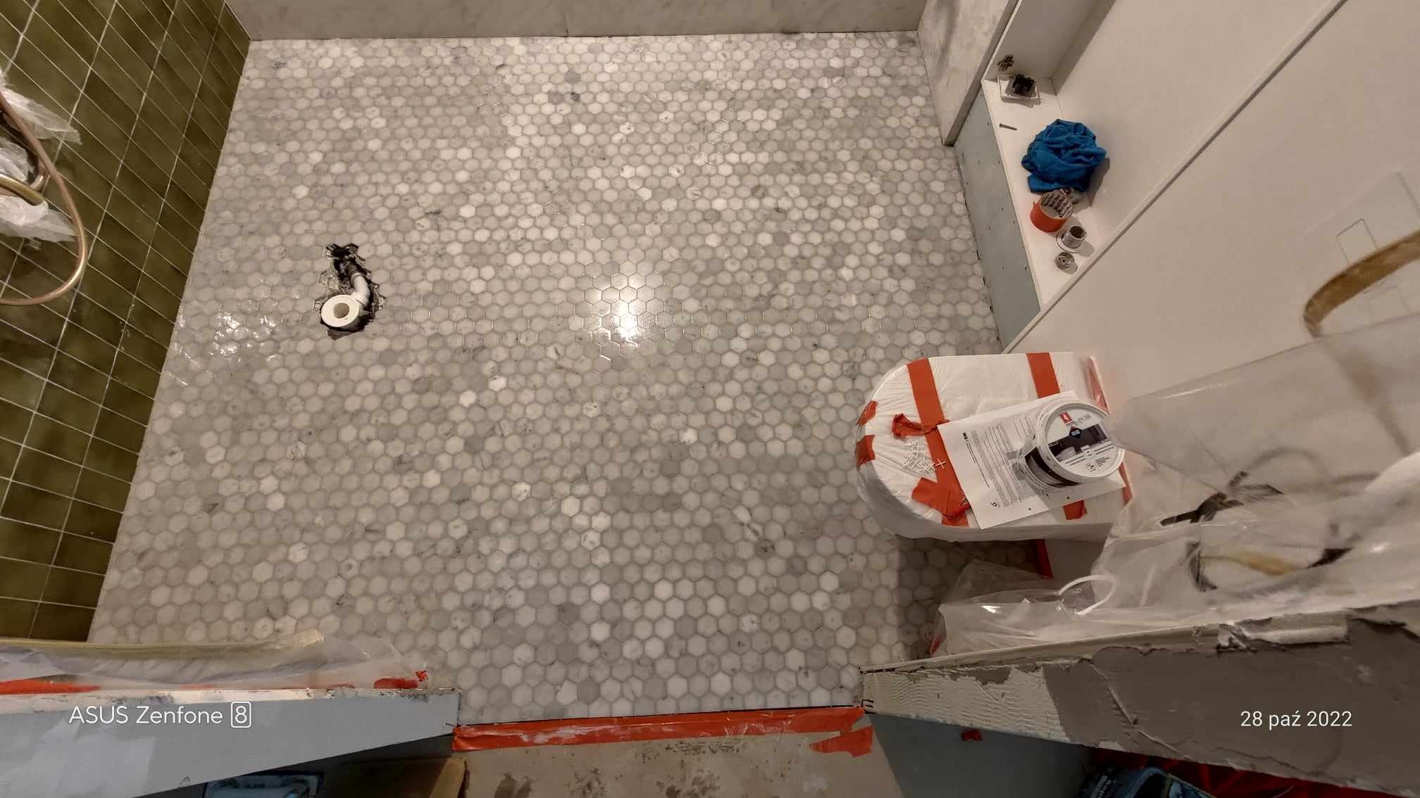 Podłoga w łazience wyłożona mozaiką z białego marmuru w kształcie plastra miodu, widoczny odpływ pod prysznic i przygotowane materiały budowlane