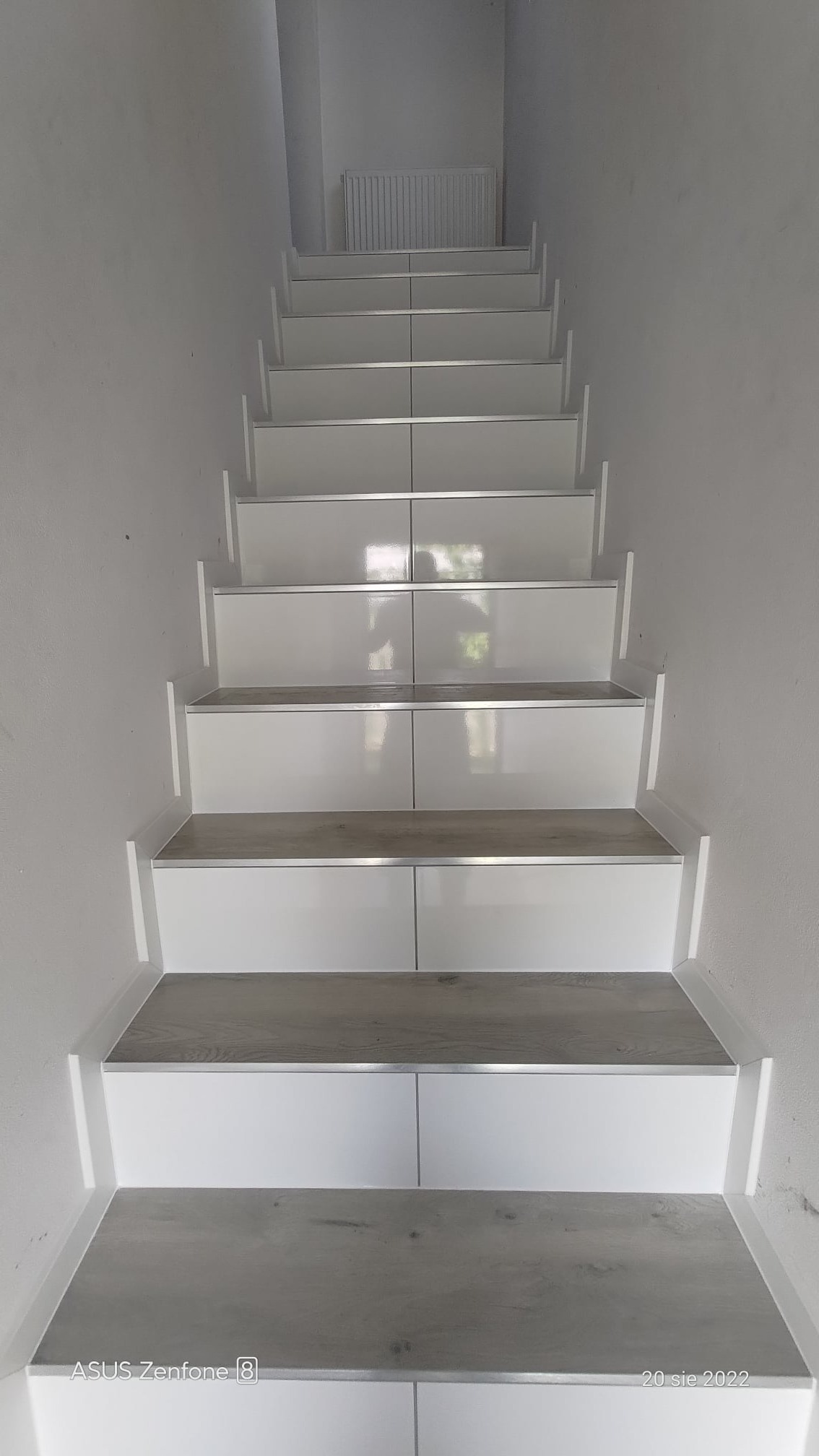 Wykończone schody z białymi podstopnicami i stopniami imitującymi drewno, zabezpieczone listwami aluminiowymi.
