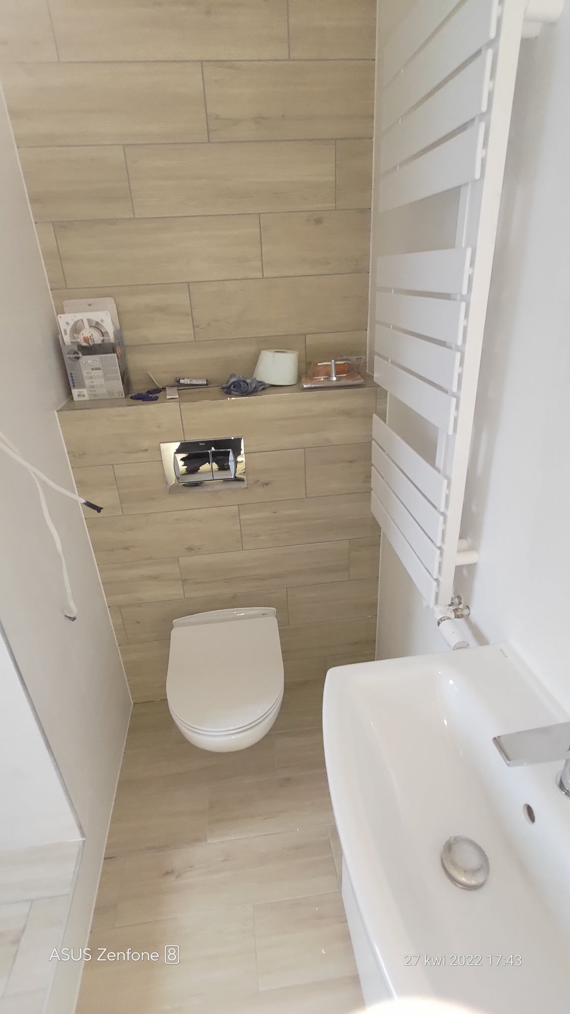 Wykończona łazienka z beżowymi płytkami imitującymi drewno, wiszącą toaletą z chromowanym przyciskiem spłukiwania, białym grzejnikiem drabinkowym i umywalką z baterią.