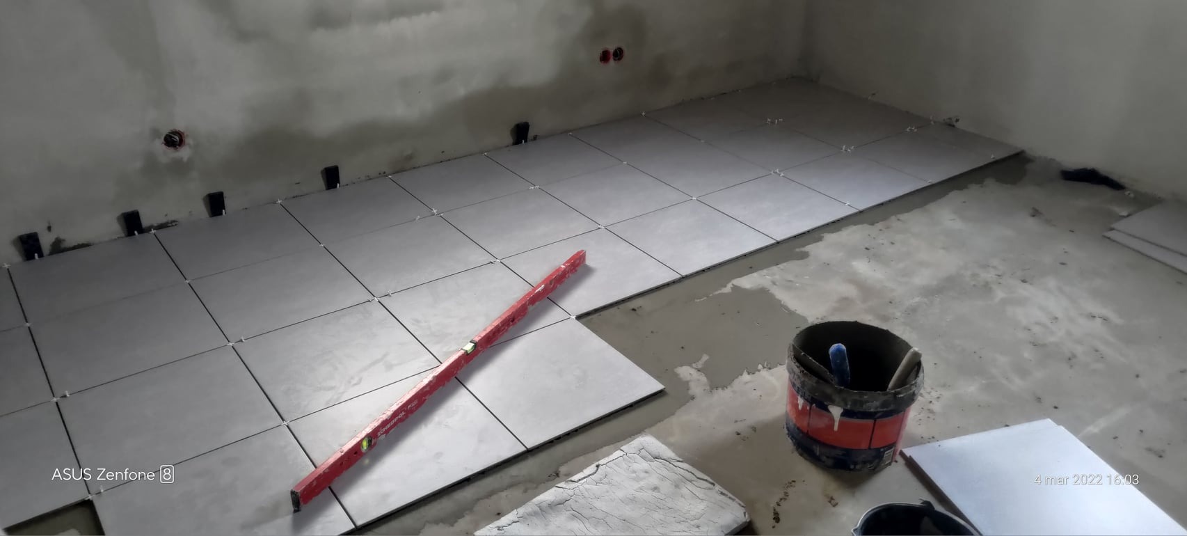 Szare płytki podłogowe ułożone częściowo w narożnym pomieszczeniu, poziomica na płytkach, wiadro z narzędziami w rogu, widoczne krzyżyki dystansowe.