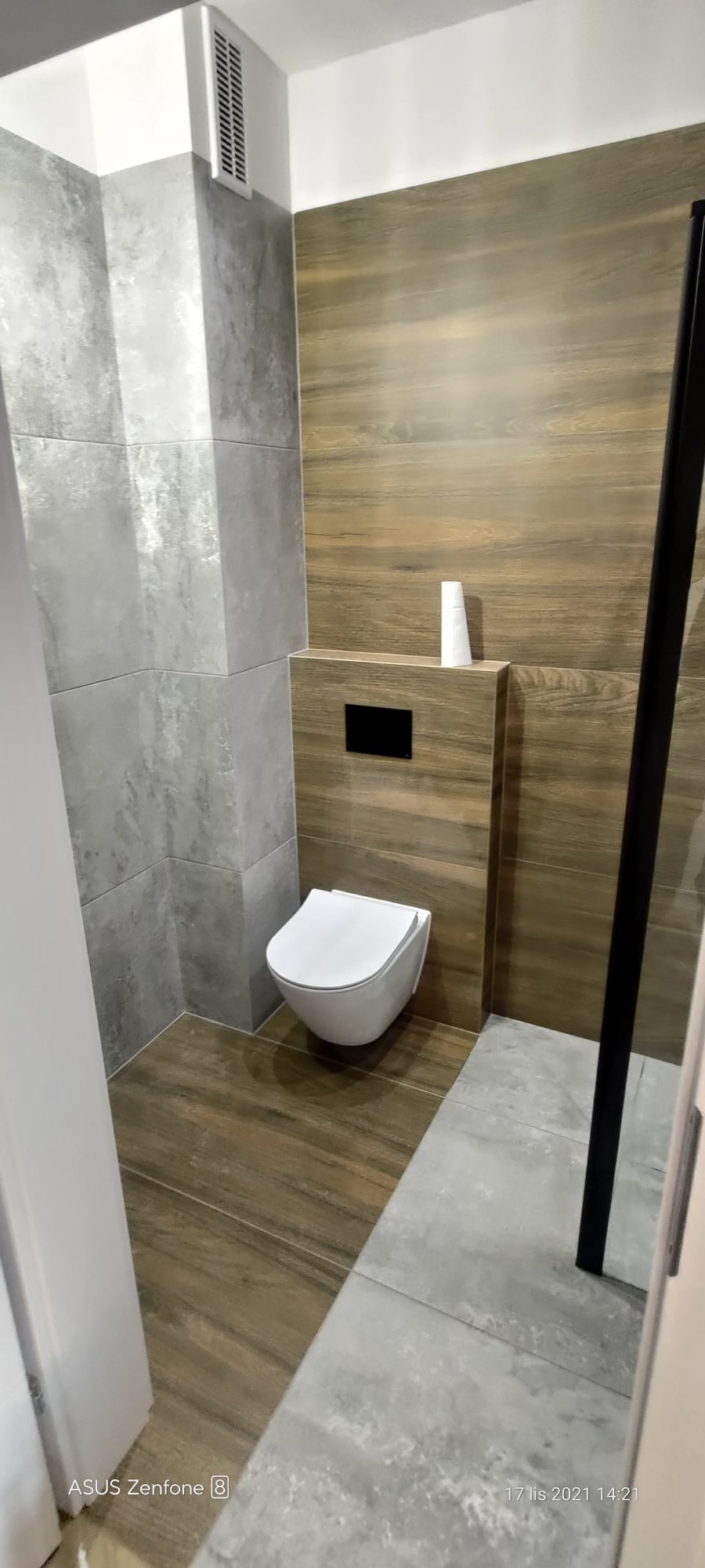 Nowoczesna toaleta podwieszana w łazience wykończonej płytkami imitującymi drewno i szarym gresem, z widocznym czarnym przyciskiem spłukiwania i minimalistyczną tubką kosmetyku na obudowie stelaża.