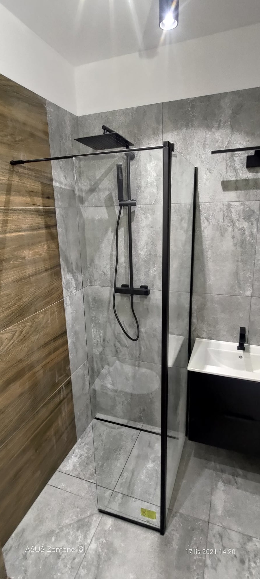 Kabina prysznicowa typu walk-in z czarnymi profilami i deszczownicą, szara płytka imitująca beton na ścianach, drewnopodobna okładzina ściany z lewej strony, czarna szafka z umywalką po prawej...