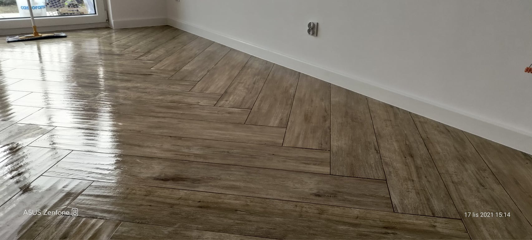 Ułożona w jodełkę, błyszcząca podłoga z płytek imitujących drewno, biała listwa przypodłogowa, widoczny mop przy oknie.