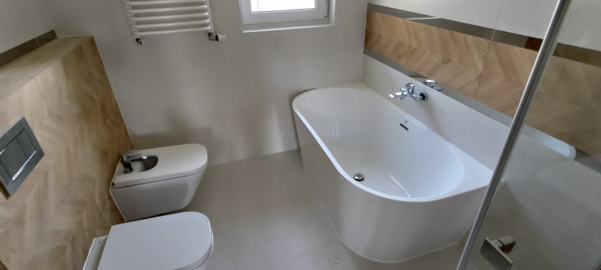 Nowoczesna łazienka z białą wanną prostokątną, wiszącą toaletą i bidetem, wykończona płytkami imitującymi drewno w strefie umywalki i toalety, z grzejnikiem drabinkowym.