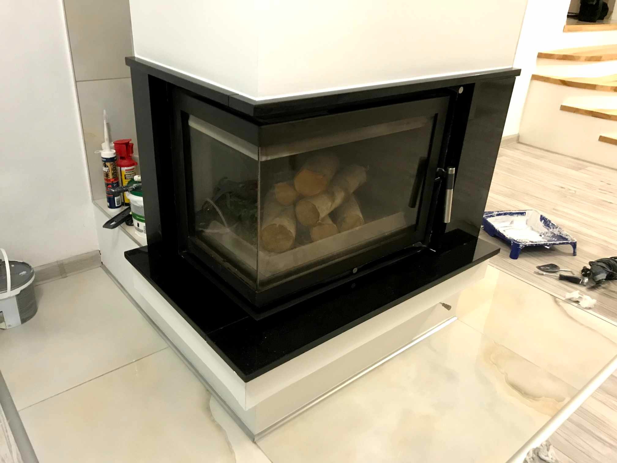Nowoczesny narożny kominek z czarną obudową i szklanymi drzwiczkami, wewnątrz ułożone drewno, obok widoczne narzędzia malarskie i chemia budowlana, podłoga wyłożona jasnymi płytkami.