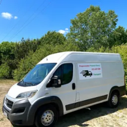 Biały bus dostawczy z naklejką reklamową firmy transportowej, zaparkowany na piaszczystym terenie z zielenią w tle, pod błękitnym niebem z pojedynczymi chmurami.