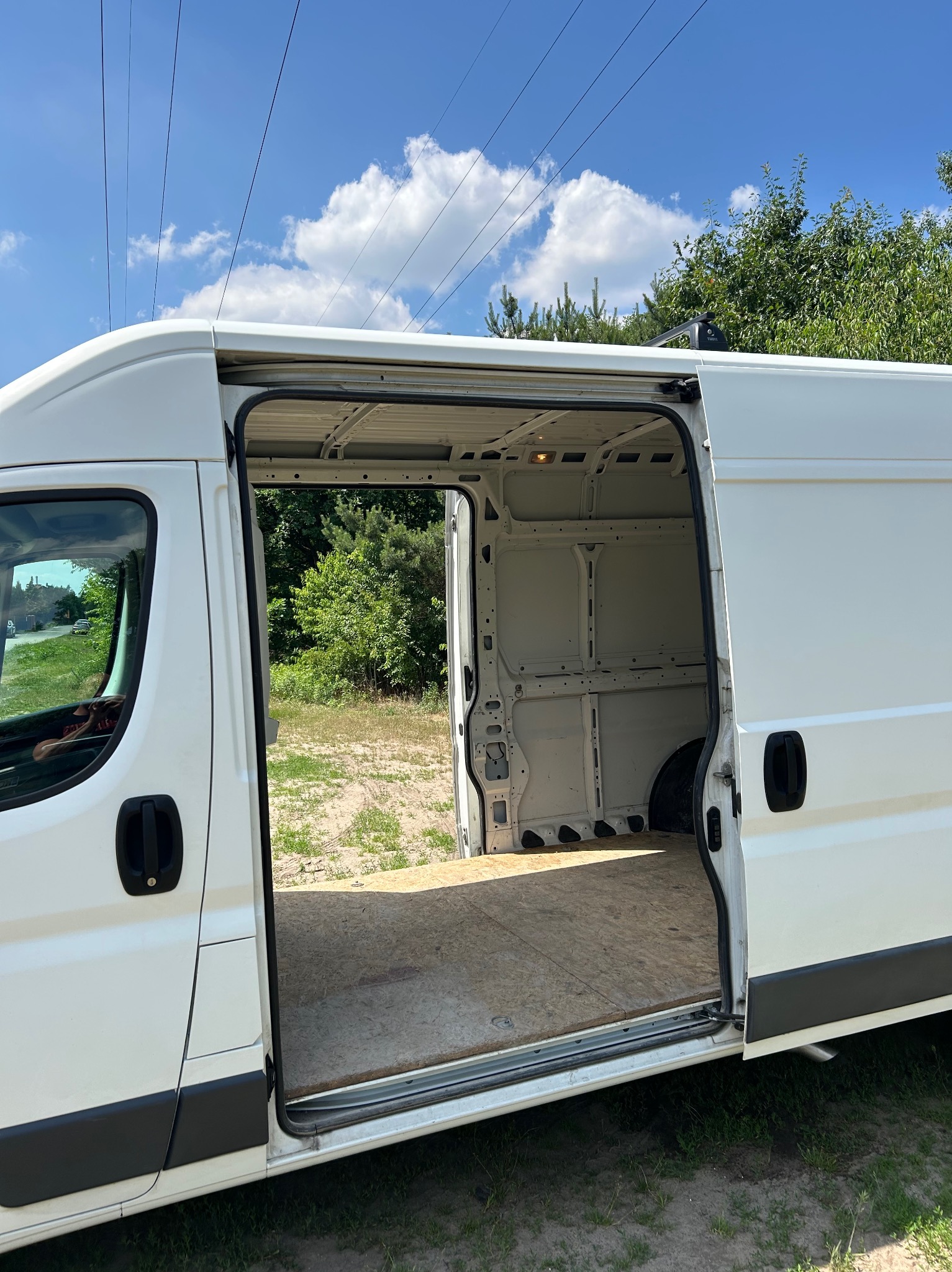 Wnętrze białego furgonetki z otwartymi drzwiami bocznymi, ukazujące drewnianą podłogę ładowni na tle zieleni drzew i błękitnego nieba.