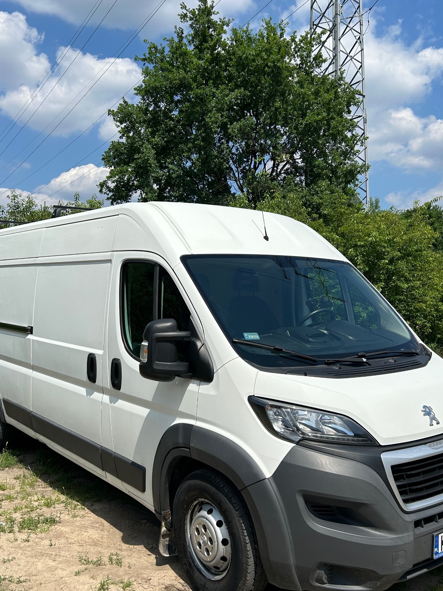 Biały samochód dostawczy Peugeot na tle zieleni i błękitnego nieba z chmurami, widoczna wieża wysokiego napięcia w tle.