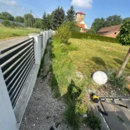 Widok na ogród z nowoczesnym ogrodzeniem panelowym, trawnikiem, kulistą lampą ogrodową i podkaszarką spalinową przy krawężniku.