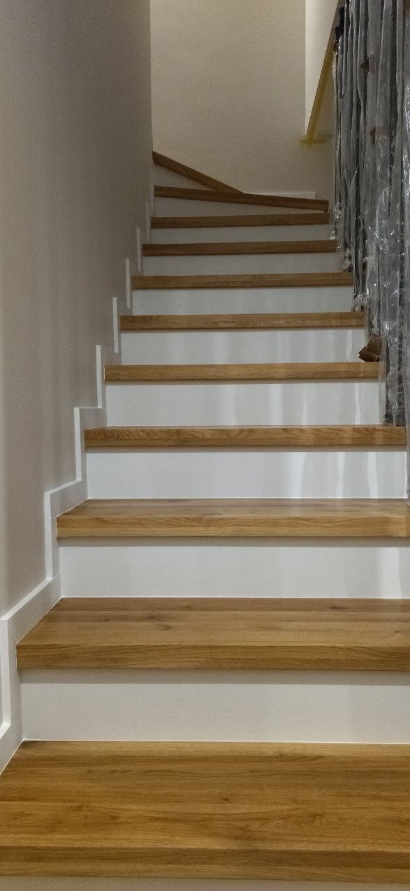 Schody z drewnianymi stopniami i białymi podstopnicami, zabezpieczone folią ochronną na balustradzie, widok z dołu.