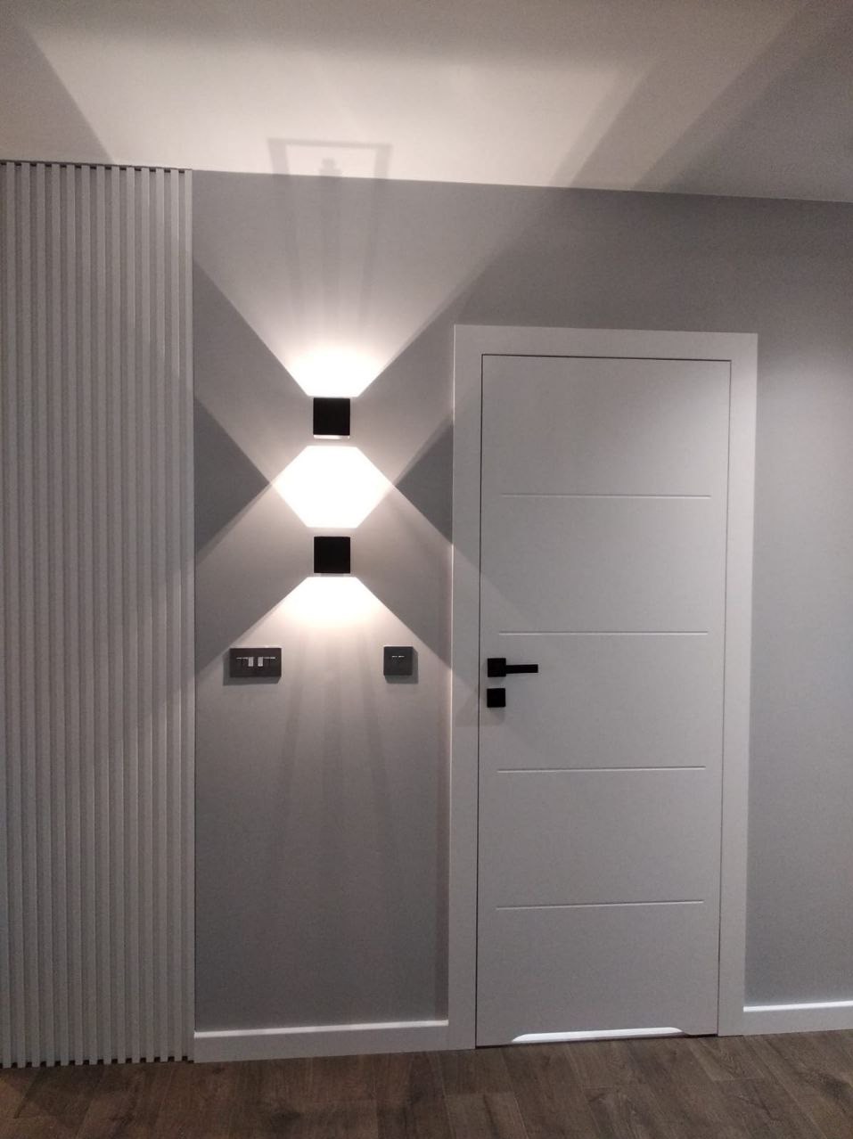Nowoczesne wnętrze: białe drzwi z czarną klamką, panel dekoracyjny z pionowymi żłobieniami i lampa ścienna z dwoma kwadratowymi kloszami, rzucająca stożkowe światło na szarą ścianę obok gniazdek.