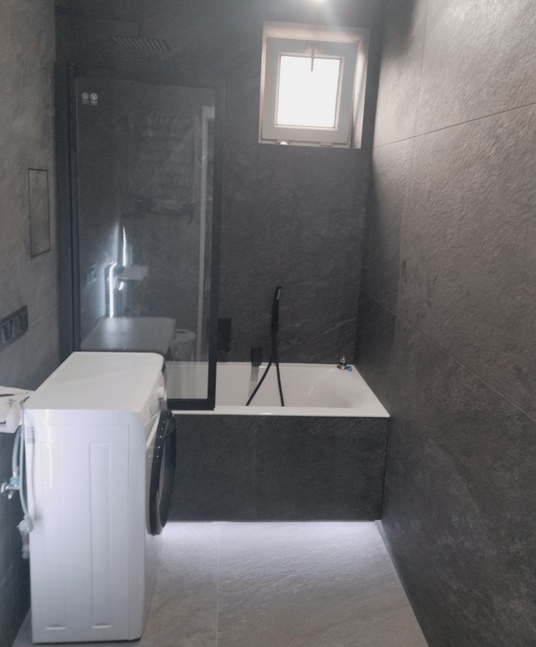 Nowoczesna łazienka w odcieniach szarości z pralką, kabiną prysznicową za szkłem i podświetlaną podłogą, widok na wannę i małe okno.