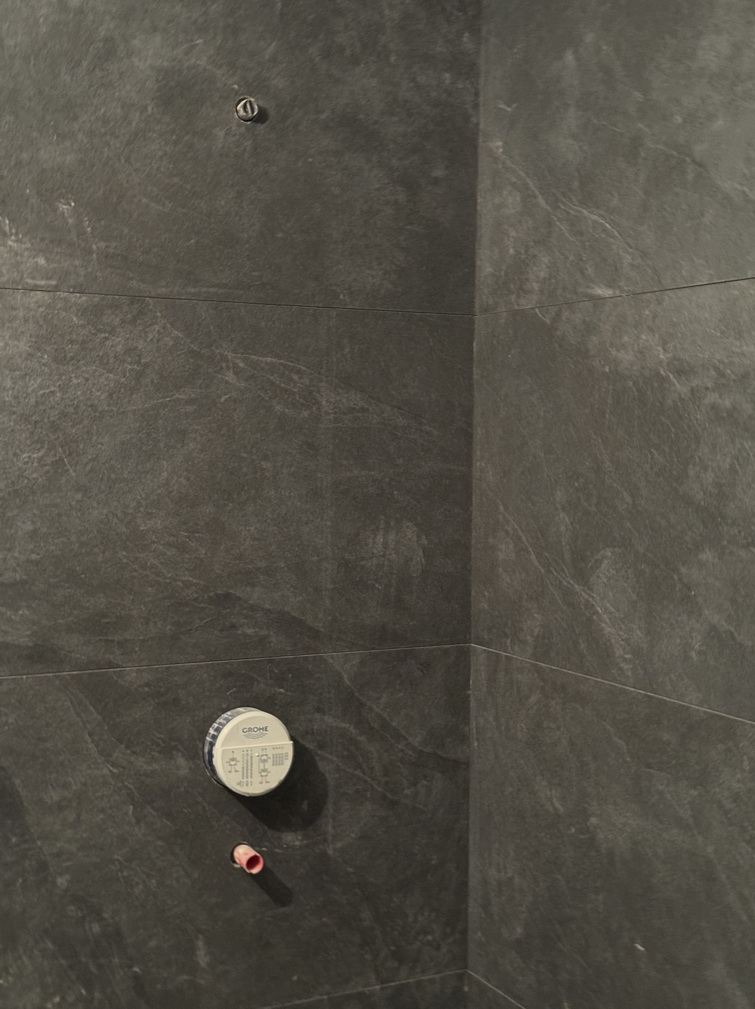 Szare płytki imitujące kamień na ścianie łazienki, widoczne przyłącza Grohe i rurka, etap prac wykończeniowych.