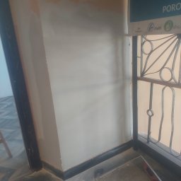 Prywatnie - Fragment ściany po szpachlowaniu, widoczne niedoskonałości przed malowaniem. Dolna część ściany z ciemną listwą przypodłogową, w tle fragment okna z kratą.