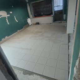 Prywatnie - Wnętrze w trakcie remontu: ułożone jasne płytki podłogowe w kwadraty, ściany częściowo wykończone, widoczne niedoskonałości i worki z materiałami budowlanymi.