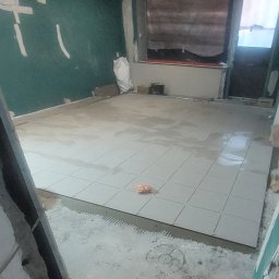 Prywatnie - Wnętrze w trakcie remontu: ułożone jasne płytki podłogowe, ściany przygotowane do malowania, widoczny worek z zaprawą. Fragment widoku na okno.