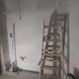 Prywatnie - Wnętrze łazienki w trakcie prac wykończeniowych. Widoczna drabina oparta o ścianę z częściowo ułożonymi płytkami, instalacja wodna i narzędzia. W tle sufit z płyt karton-gips.
