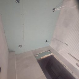 Prywatnie - Montaż sufitu z płyty GK w łazience, widoczne otwory na oświetlenie i wystające kable. Płytki na ścianach, przygotowanie do wykończenia.