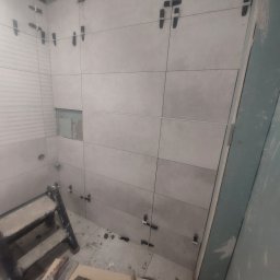 Prywatnie - Ściana łazienki w trakcie wykańczania z szarymi płytkami, narzędziami i drabinką, widoczne klipsy poziomujące płytki.