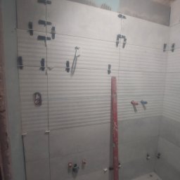 Prywatnie - Układanie jasnych, poziomych płytek z fakturą w łazience. Widoczne krzyżyki dystansowe, poziomica i fragment instalacji. W tle surowy fragment ściany.