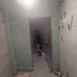 Prywatnie - Remont łazienki: świeżo położone płytki na ścianie, widoczne narzędzia i materiały budowlane, surowa ściana w tle. Prace w toku, widok na sąsiednie pomieszczenie.