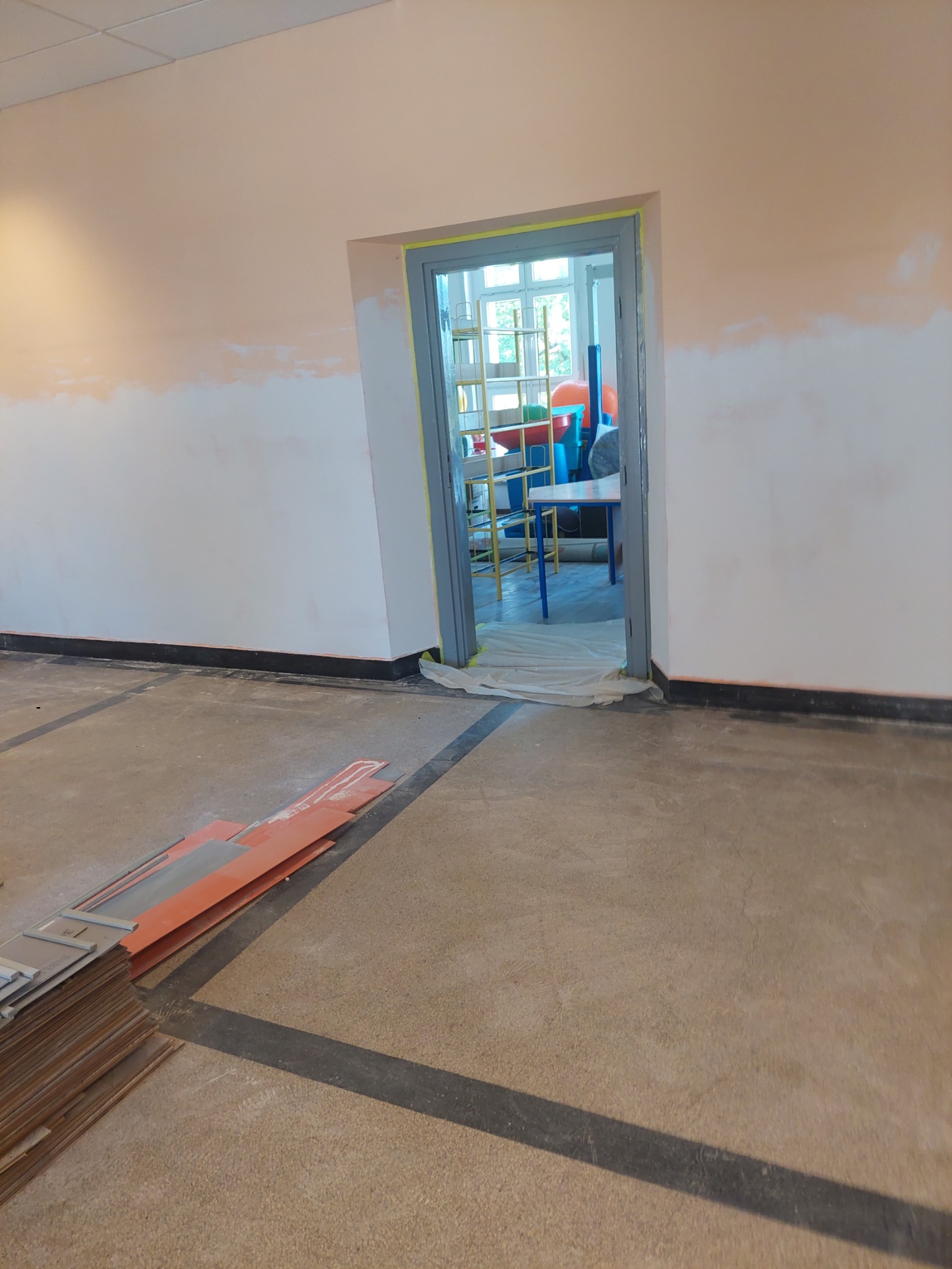 Przygotowanie do malowania ścian: fragment pomalowanej ściany, zabezpieczone futryny drzwiowe taśmą malarską, panele podłogowe i folia ochronna na podłodze.