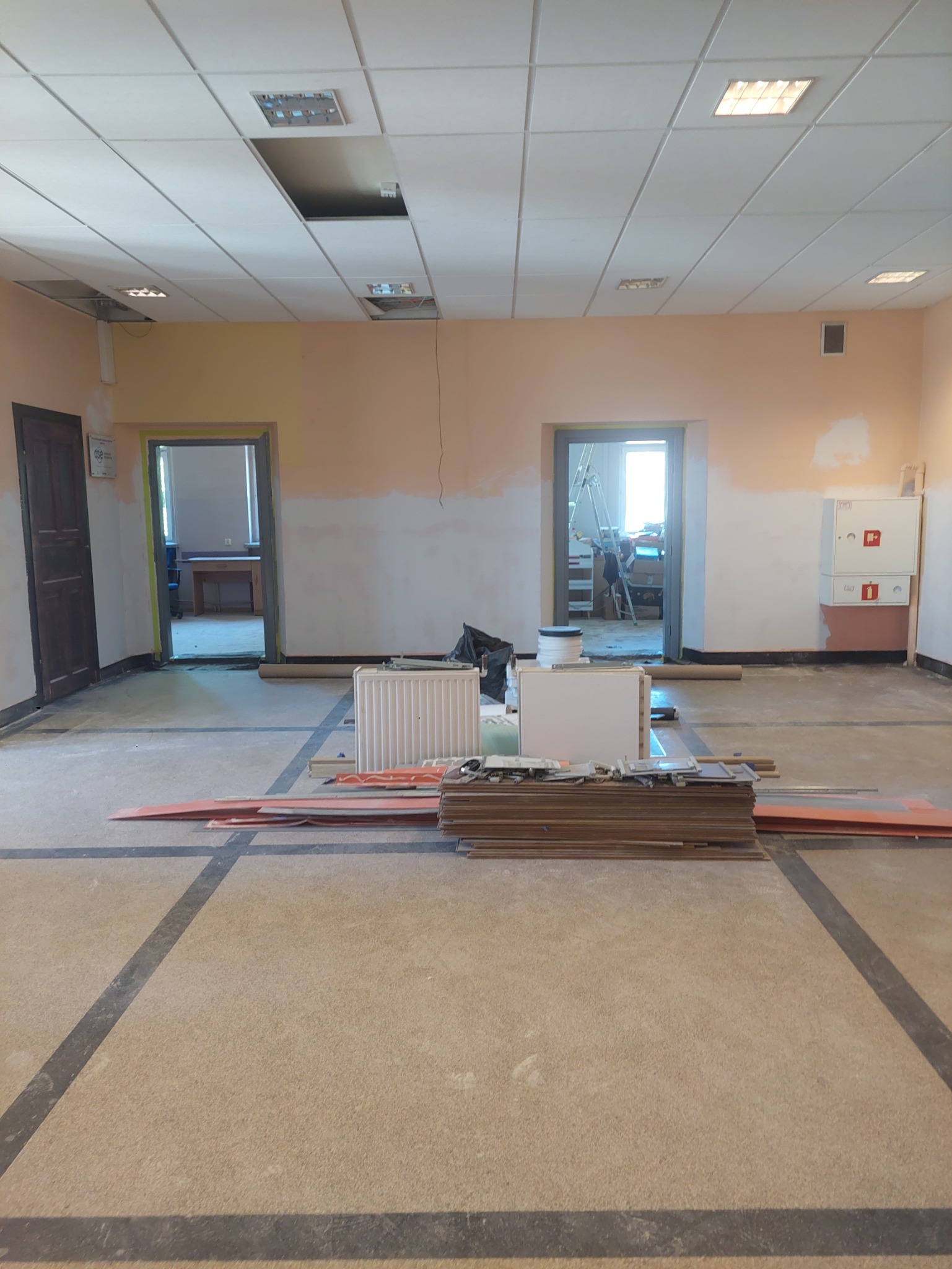 Wnętrze w trakcie remontu: przygotowane płyty, grzejnik, ściany częściowo pomalowane, sufit z brakującymi panelami. Widoczne drzwi i okna.