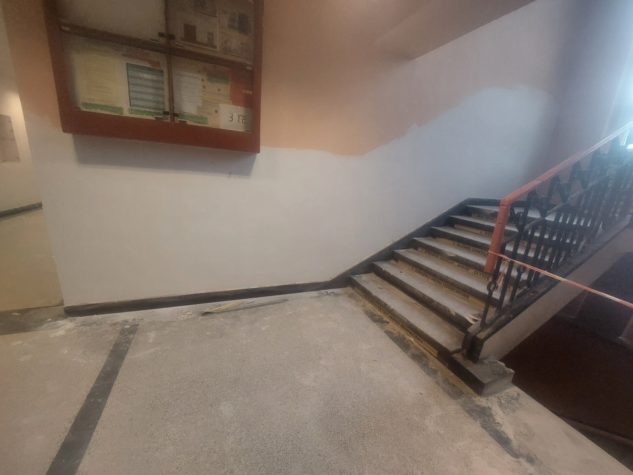 Klatka schodowa w trakcie remontu: ściany częściowo pomalowane, schody z napisami, widoczny fragment tablicy informacyjnej. Prace wykończeniowe w budynku.