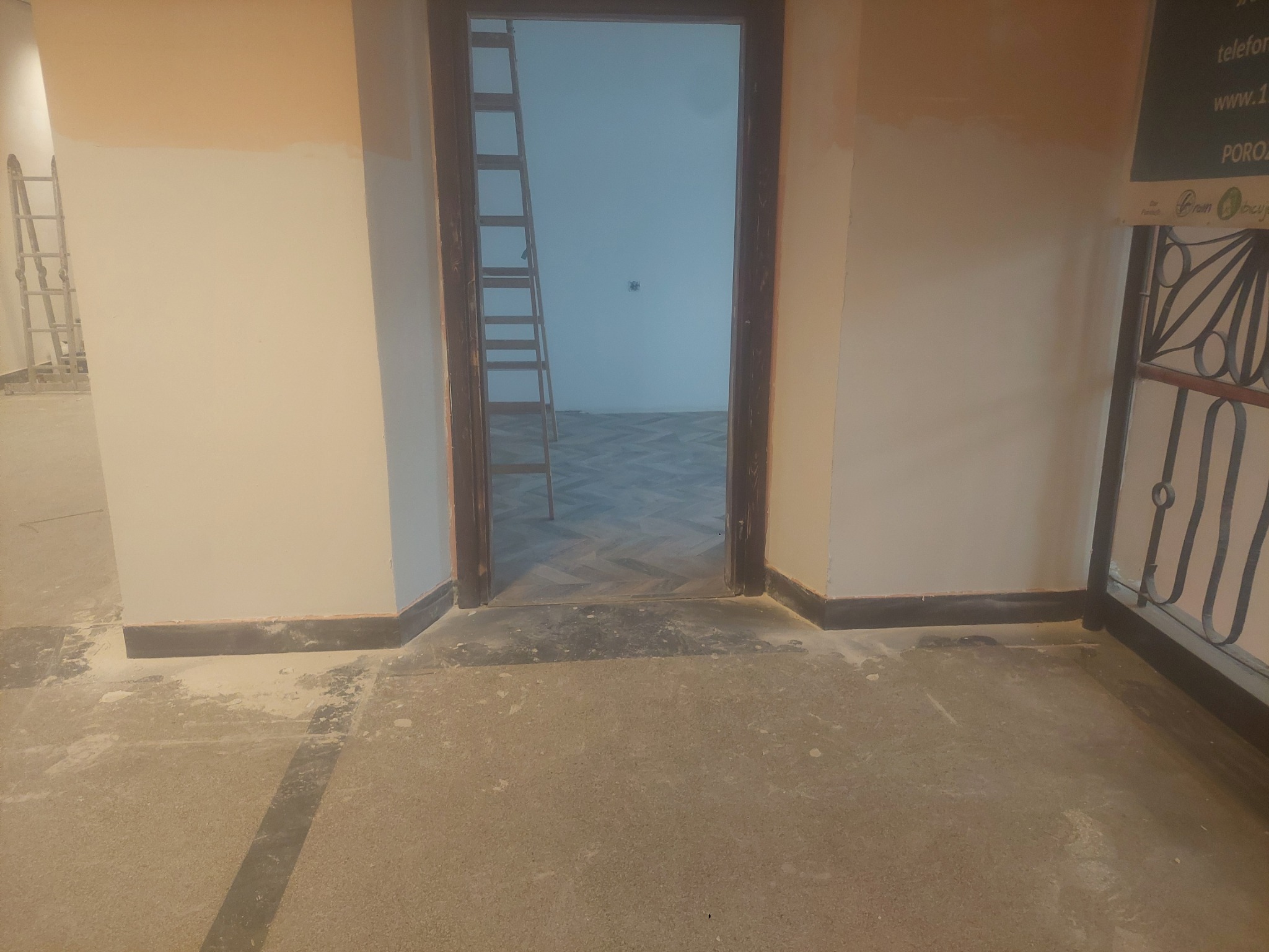 Wnętrze w trakcie remontu: widoczne ślady po pracach malarskich na ścianach, podłoga z resztkami materiałów budowlanych, drabina w tle. Widok na sąsiednie pomieszczenie.