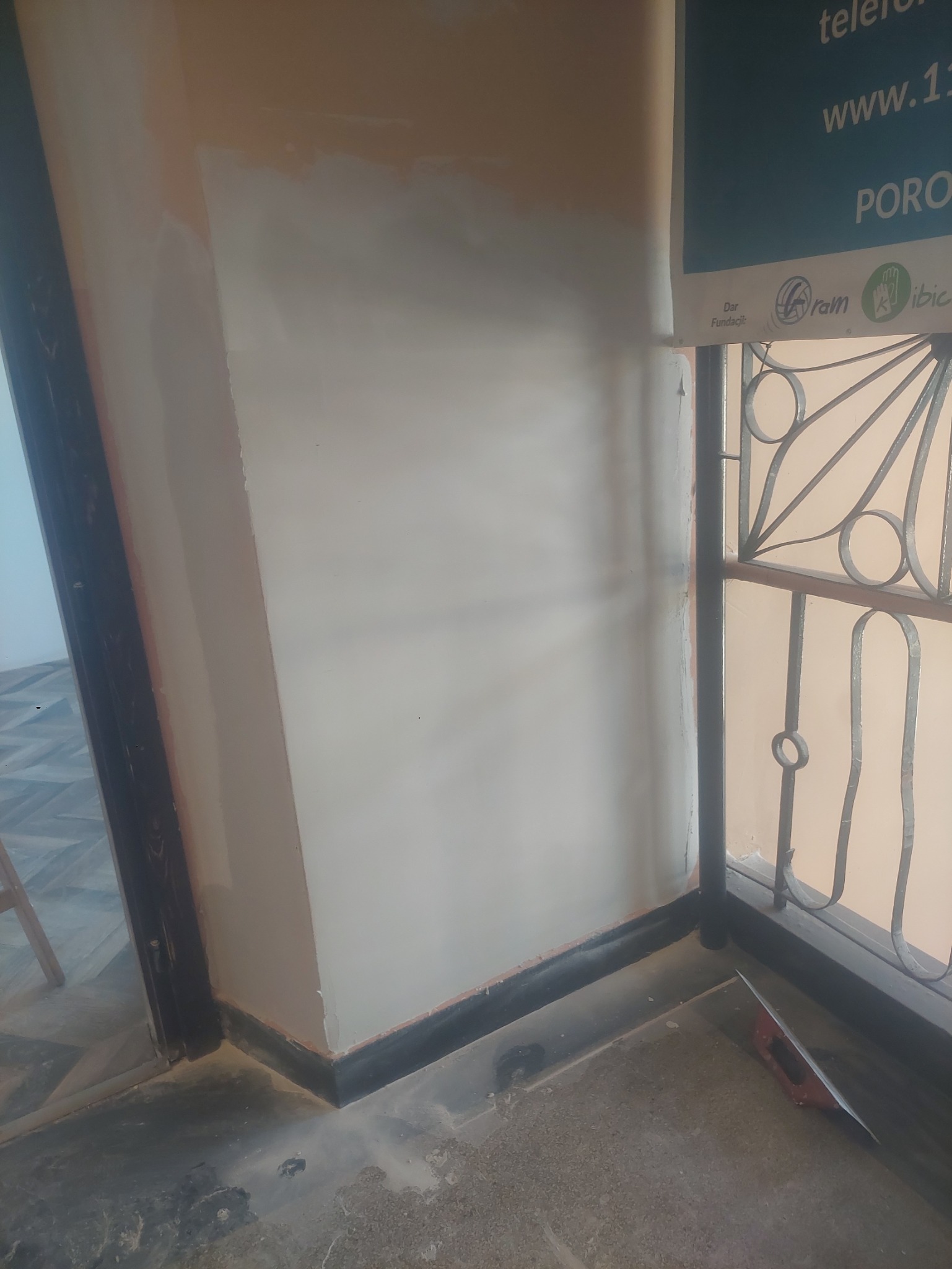 Fragment ściany po szpachlowaniu, widoczne niedoskonałości przed malowaniem. Dolna część ściany z ciemną listwą przypodłogową, w tle fragment okna z kratą.